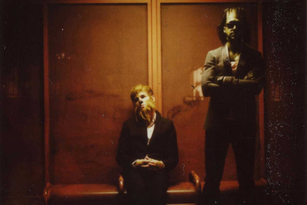 Promotional portrait of indie rock duo Dead Man's Bones, Ryan Gosling and Zach Shields, posing in warm ambient lighting.